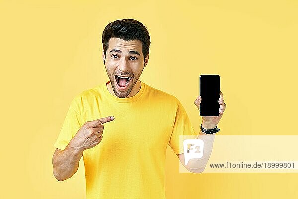 Aufgeregter schreiender Mann  der mit dem Finger auf sein Handy zeigt und in die Kamera schaut  auf gelbem Studiohintergrund