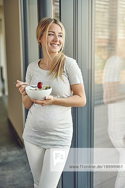 Glückliche  gesunde  schwangere  junge  blonde Frau  die an ein Terrassenfenster gelehnt steht und lächelnd nach draußen schaut  während sie eine Schüssel mit frischem Obstsalat genießt