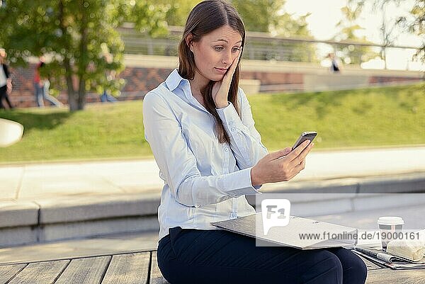 Attraktive schlanke junge Frau  die im Freien in einem Stadtpark sitzt und mit besorgtem Blick und der Hand im Gesicht eine SMS auf ihrem Handy liest