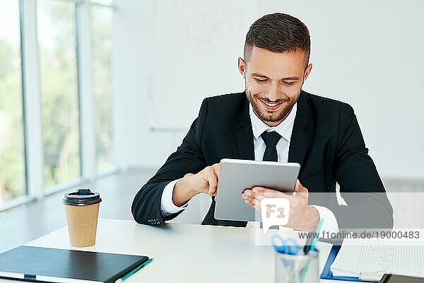 Glücklicher erfolgreicher Geschäftsmann in eleganter Suite mit Tablet in modernem kreativen Büro sitzend. Geschäftskonzept