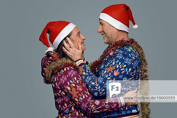 Festliche romantische Paar feiert Weihnachten in Santa Hats und bunten Xmas themed Kleidung umarmt einander mit glücklichen Lächeln in einer Seitenansicht vor einem grauen Hintergrund