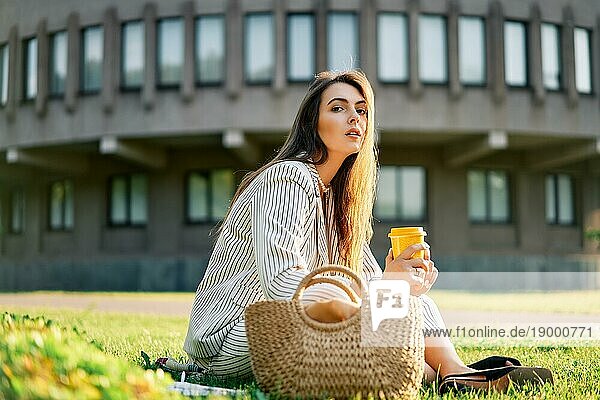 Junge stilvolle Frau trinkt Kaffee und entspannen Sie sich auf einem Rasen im Stadtpark sitzen. Pause Zeit Konzept
