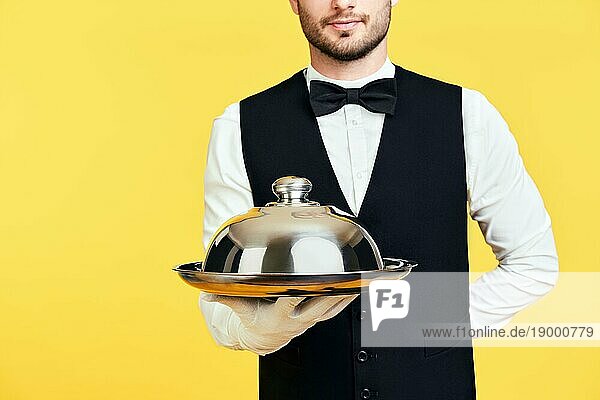 Junge gut aussehende Kellner hält Metalltablett mit Deckel bereit zu dienen auf gelbem Hintergrund