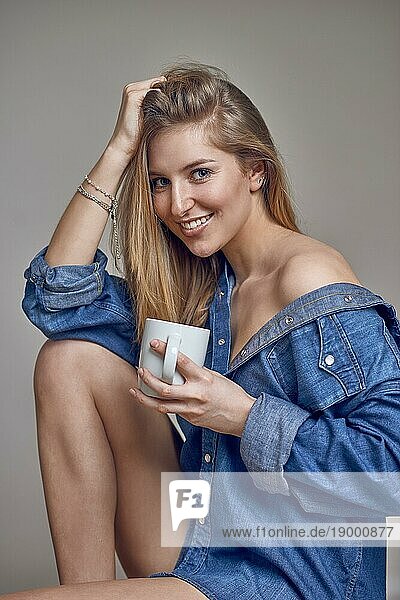 Attraktive  trendige  junge  blonde Frau mit Jeanshemd  das über die Schulter geöffnet ist  und nackten Beinen  die mit einer Tasse Kaffee in der Hand sitzt und fröhlich in die Kamera lächelt  während sie sich die Haare hält