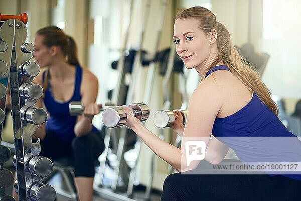 Fitte  gesunde Frau  die vor einem großen Wandspiegel in einem Fitnessstudio mit Gewichten trainiert und sich umdreht  um über ihre Schulter in die Kamera zu schauen  während sie zwei Hanteln hält