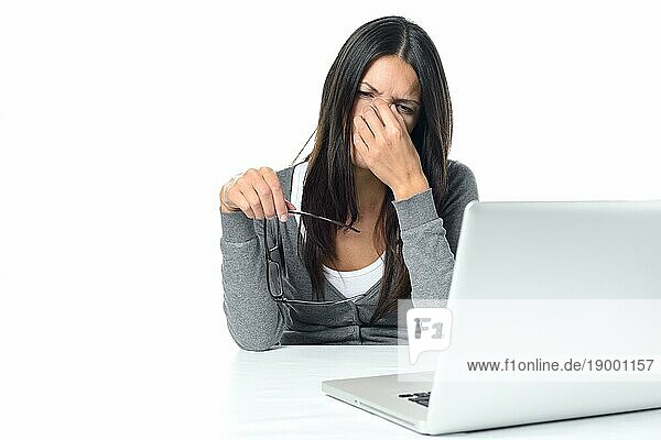 Junge Frau  die unter einer Überanstrengung der Augen an ihrem Laptop leidet  nimmt ihre Brille ab und reibt sich mit schmerzverzerrtem Gesichtsausdruck die Augen mit ihrem Finger  auf weißem Hintergrund