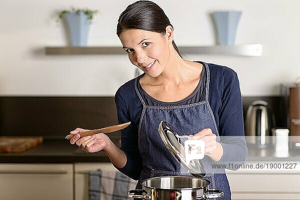 Junge Frau  die das Essen für das Abendessen auf dem Herd in ihrer Küche kocht und dabei den Deckel eines Edelstahltopfes und eine Holzkelle hält  während sie in die Kamera lächelt