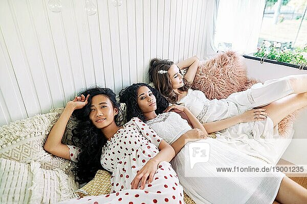 Schöne Multi ethnische Mädchen haben Spaß zusammen  entspannen und liegen auf dem Bett im Inneren des Wohnmobils. Frauen Freundschaft und Frauen Schönheit Konzept. Kaukasisch  afrikanisch amerikanischen und asiatischen Mädchen posieren bei Licht Innen