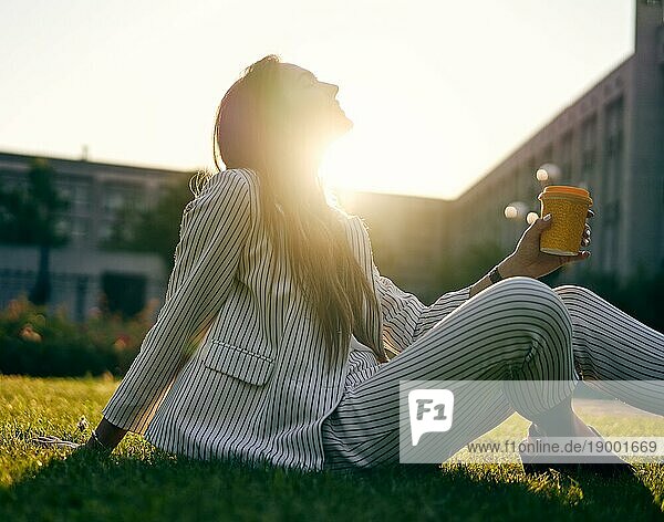 Junge stilvolle Frau trinken Kaffee und entspannen Sie sich auf Stadtpark Gras im Sonnenschein Licht sitzen. Pause Zeit Konzept