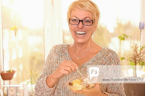 Lebhafte attraktive Frau mittleren Alters mit Brille  die eine gesunde Schüssel vitaminreichen Obstsalat ißt und mit einem freundlichen Lächeln voller Lebensfreude in die Kamera schaut