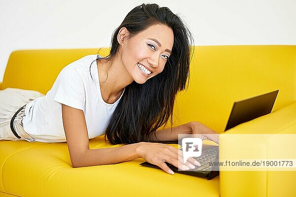 Hübsche lächelnde Frau mit Laptop auf einer gemütlichen gelben Couch auf weißem Hintergrund. Technologie  Freiberufler Konzept