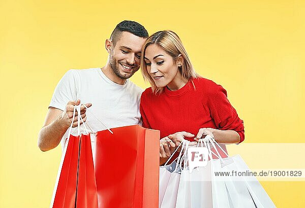 Junges glückliches Paar  das Einkaufstaschen in den Händen hält und ihren Einkauf vorgelbem Hintergrund betrachtet