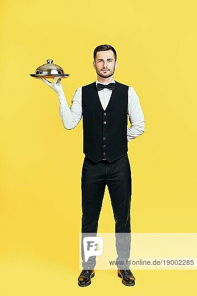 Full Länge Porträt der eleganten gut aussehend Kellner hält Tablett mit Deckel bereit  auf gelbem Hintergrund zu dienen. Restaurant  Horeca Konzept