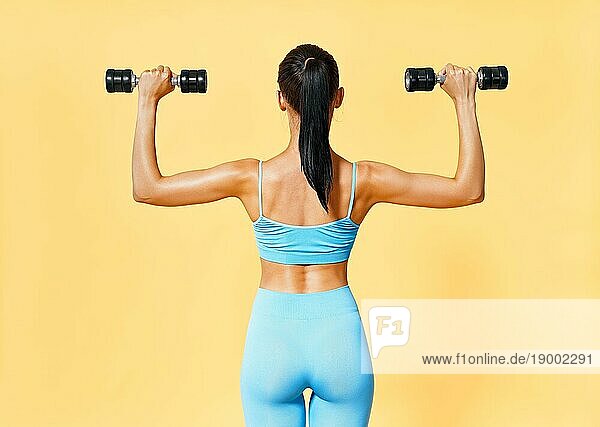 Rückenansicht einer sportlichen Frau in Sportkleidung beim Training mit Hanteln auf gelbem Hintergrund. Stärke und Motivation Konzept