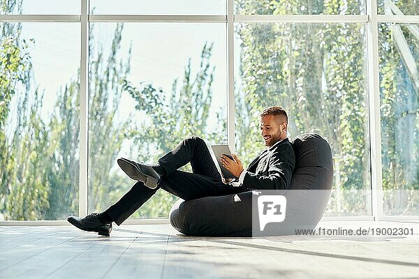 Lächelnde erfolgreiche Geschäftsmann in eleganten Suite mit Tablet entspannen auf Sitzsack Stuhl mit Panoramafenstern Hintergrund in modernen kreativen Büro