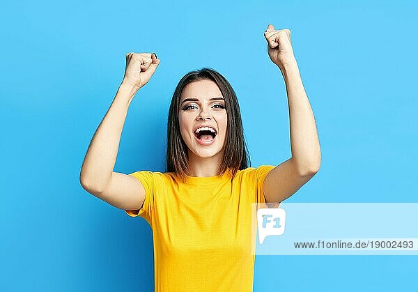 Glücklich aufgeregt junge Frau feiern Erfolg mit den Händen nach oben vorblauem Hintergrund. Sieg  Emotionen Konzept