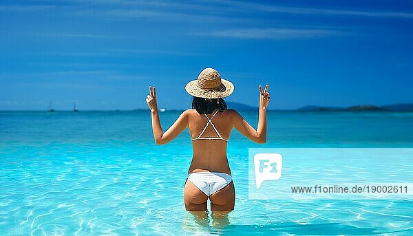 Glückliche sexy Frau in weißem Bikini und Strohhut zeigt Friedenszeichen mit beiden Händen entspannen in türkisfarbenen Meerwasser auf tropischen Strand im Paradies Insel. Entspannen  Urlaub  Sommer Reise Konzept