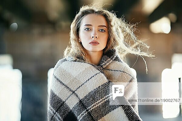 Close up Porträt der schönen jungen Frau in Wolldecke eingewickelt Blick zur Kamera auf windigen sonnigen Tag auf städtischen Straße Hintergrund. Ausdruck  weibliche Schönheit  Entspannung  Komfort Konzept