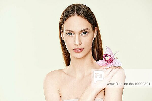 Close up Porträt der schönen jungen Frau mit natürlichen Make up und Orchidee in der Hand Blick in die Kamera auf Studio Hintergrund. Weibliche Schönheit  Spa  saubere Haut Konzept