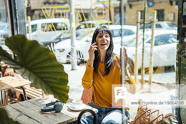 Happy mature woman waving and talking on smart phone in cafe
