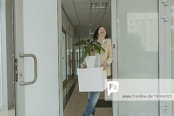 Cheerful senior businesswoman holding box with potted plant in doorway at office
