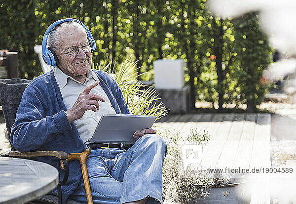 Thoughtful senior man wearing wireless headphones sitting with tablet PC