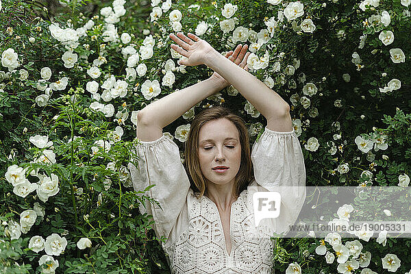 Young woman with eyes closed and arms raised by white flower bush