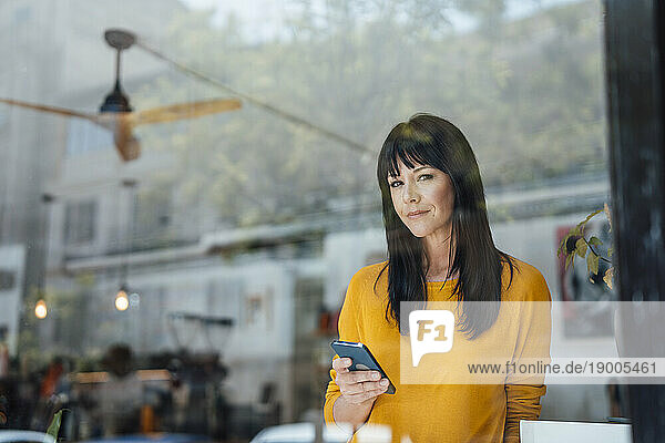 Smiling woman with smart phone seen through glass in cafe