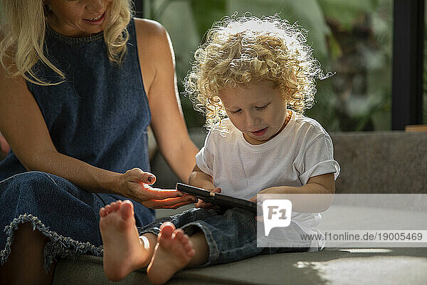 Son with curly hair using tablet PC sitting next to mother at home