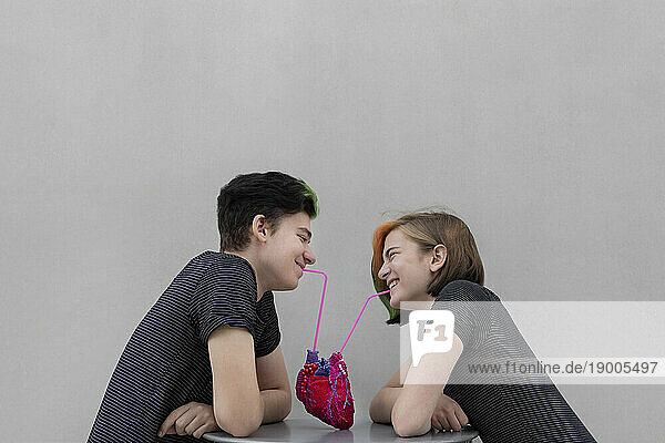 Cheerful teenage couple drinking from heart against gray background