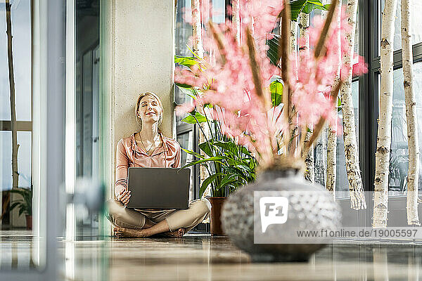 Smiling businesswoman sitting with laptop leaning on column