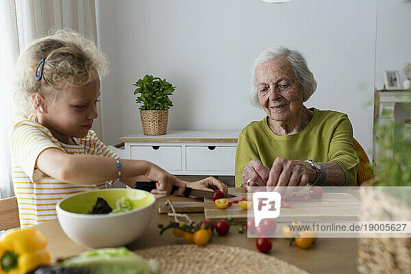 Smiling grandmother and granddaughter cutting vegetables on table