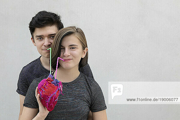 Smiling teenage couple drinking together from model heart against gray background