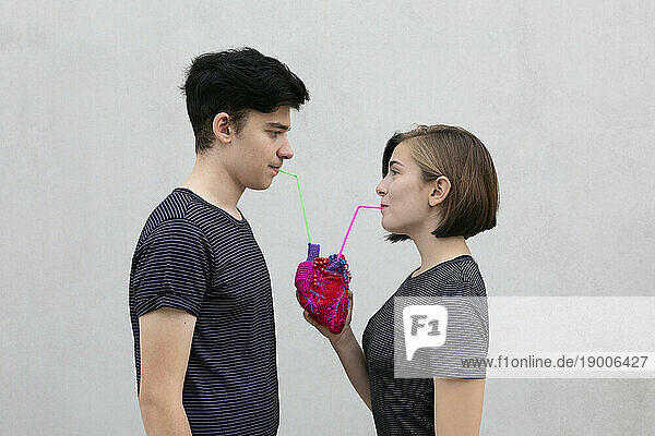 Teenage couple drinking together from model heart against gray background