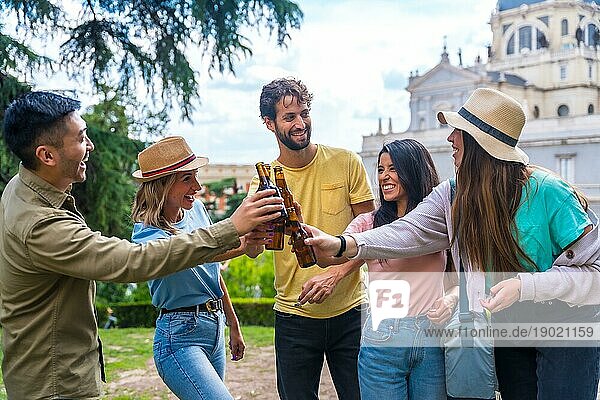 Eine multiethnische Gruppe von Freunden feiert in einem Stadtpark mit Bier. Lächelnd und mit den Flaschen anstoßend