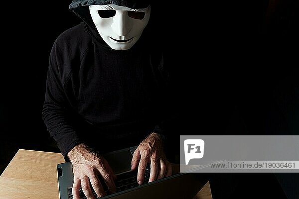 Schwarz gekleideter Hacker mit Kapuze und Maske tippt auf einem Computer CybercrimeKonzept