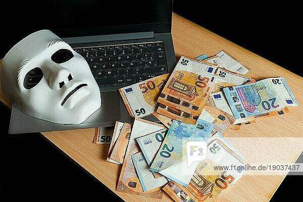 Auf einem Laptop befinden sich eine weiße Maske und ein Stapel Eurobanknoten CybercrimeKonzept