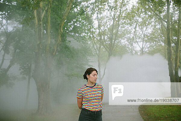 Young woman with downcast eyes standing on a path in a foggy park