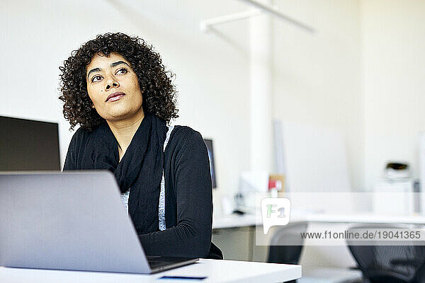 Thoughtful businesswoman with curly hair standing at desk against wall in office