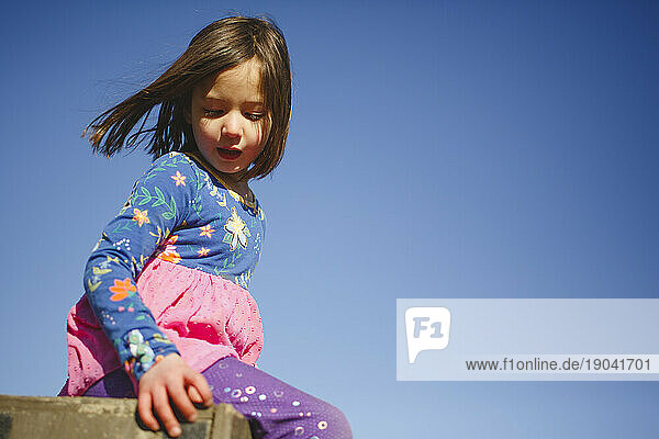 A little girl sits peacefully on a box against blue sky wind in hair