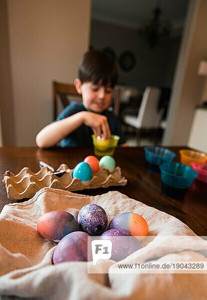 Pile of colored Easter eggs with boy dying an egg in the background.