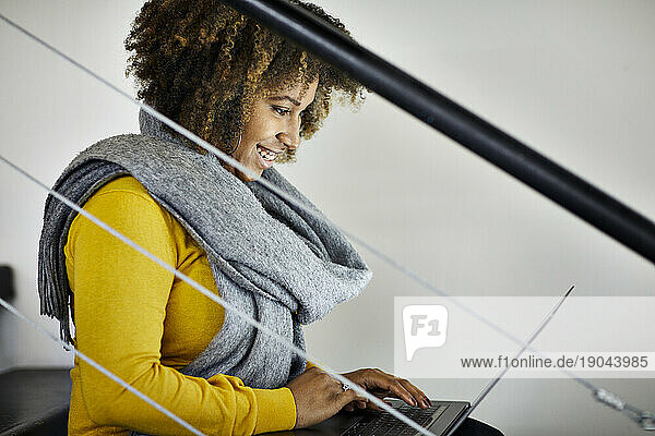 Side view of smiling businesswoman with curly hair using laptop computer while sitting on steps in office