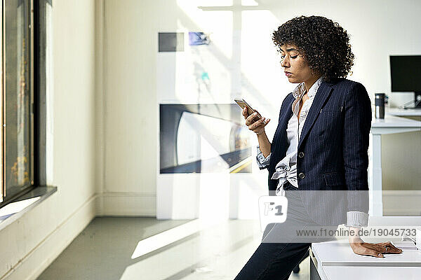 Confident businesswoman using mobile phone while standing by desk against wall in office