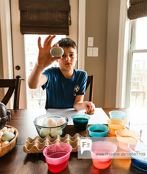 Boy holding colored Easter egg at the table with containers of dye..