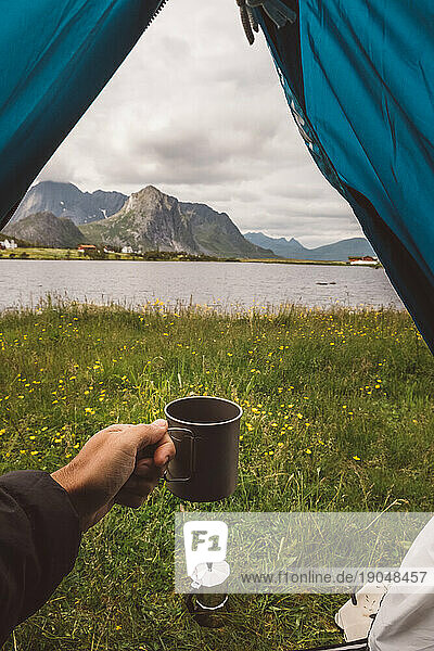 Man's hand holding a mug in a camping tent with a lake view in Norway