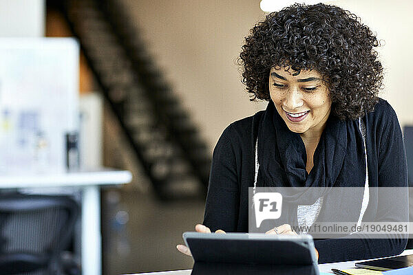 Smiling businesswoman with curly hair using tablet computer on desk in office