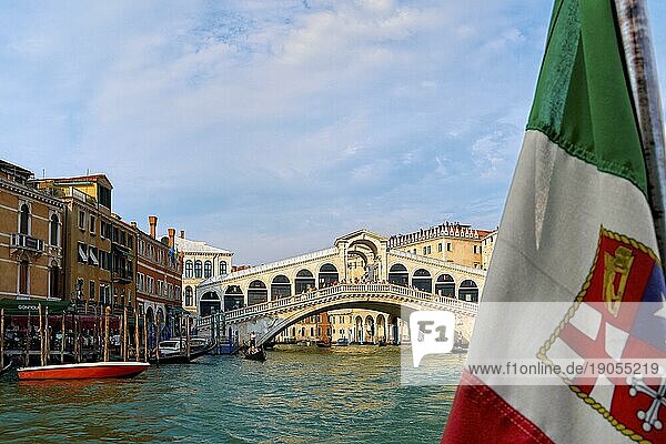 Schöner Blick auf die italienische Seeflagge und die Rialtobrücke oder Ponte Rialto am Canal Grande in Venedig  Italien. Gondel Service Station auf der linken Seite. UNESCO Weltkulturerbe Stadt. Tageslicht  Sonnenschein  weiche Wolken. Selektiver Fokus