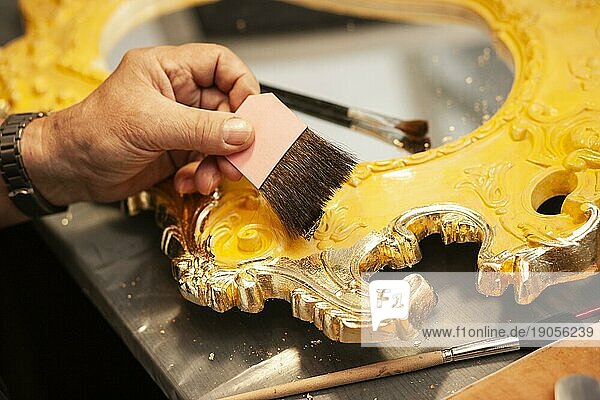 Detail eines mit Blattgold vergoldeten Spiegels  ein Mann legt ein Stück Blatgold mit einer Pinzette ab und streicht mit einem Pinsel darüber