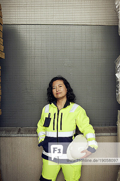 Porträt einer selbstbewussten Arbeiterin in reflektierender Kleidung  die in einer Fabrik an der Wand steht