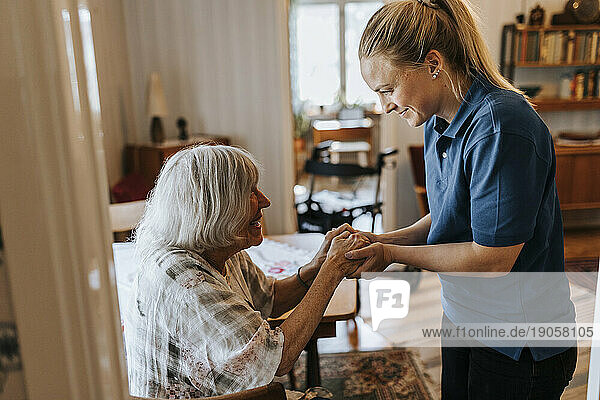 Lächelnde Mitarbeiterin im Gesundheitswesen hält die Hände einer älteren Frau zu Hause